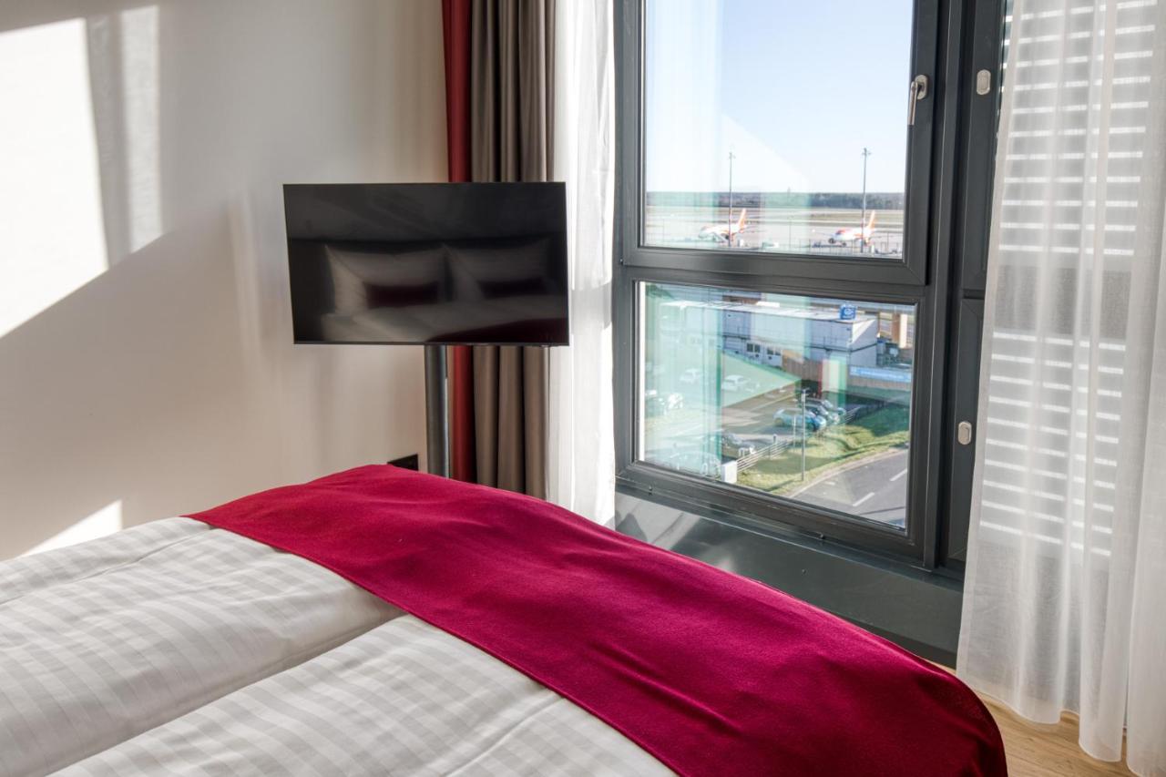 Intercityhotel Berlin Airport Ber Terminal 1 & 2 Schönefeld Exterior foto The photo shows a modern hotel room with a large bed covered in white linens and a burgundy throw blanket. In the background, there is a large window that lets in natural light, offering a view of the outside. A flat-screen TV is mounted on the wall 