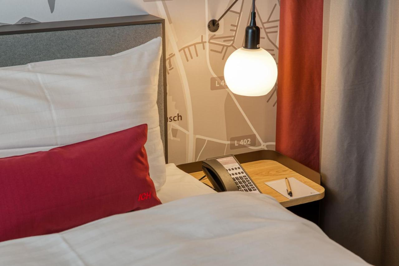 Intercityhotel Berlin Airport Ber Terminal 1 & 2 Schönefeld Exterior foto The photo shows a cozy bedroom setup. There's a neatly made bed with white bedding and a red decorative pillow. Beside the bed, a small bedside table holds a telephone and a notebook or notepad, along with a pen. A round pendant light hangs above the